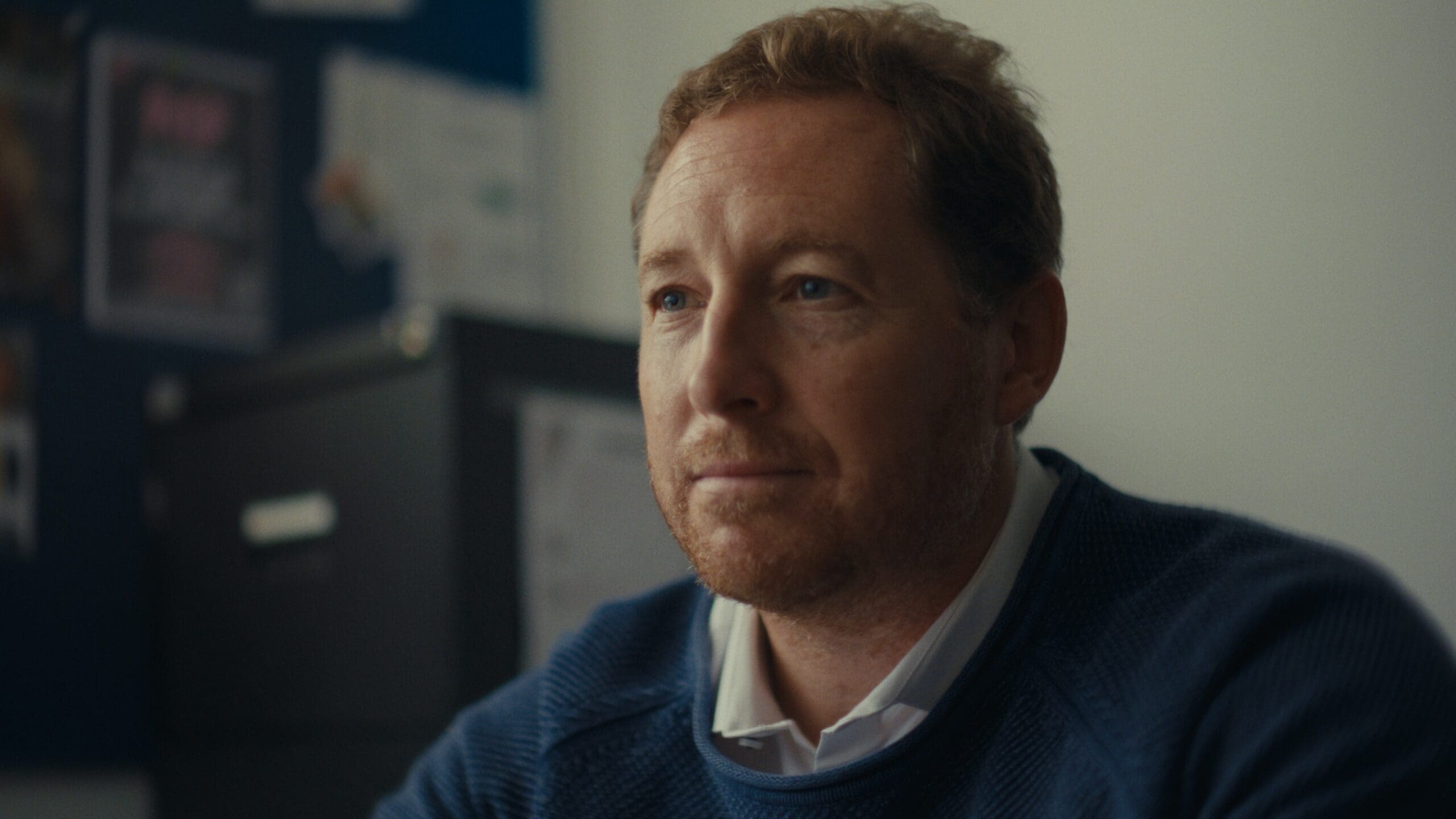 Head and shoulders if a white man wearing a warm jumper, sat in what could be an office. He looks tired but hopeful.