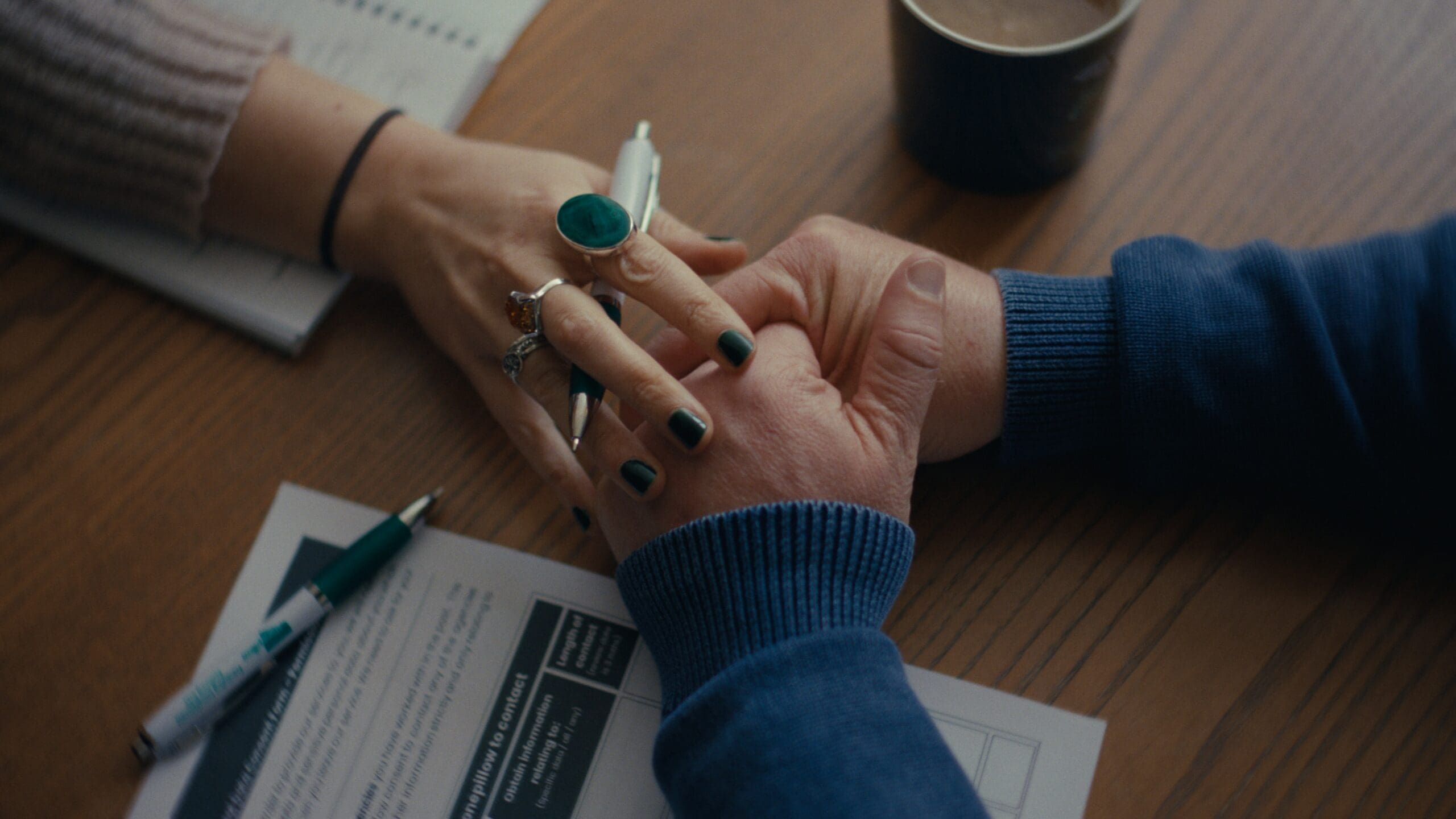 Close-up shot of someone's hands clasped in worry, leaning on a table. There are official-looking forms and a pen next to them. Another person's hand is reaching out to give comfort - it appears they have broken off in the middle of taking notes.