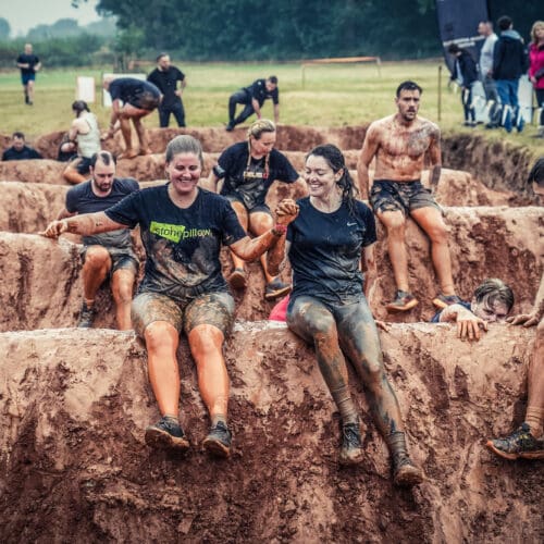 Two young women caked in mud sit at the top of a muddy bank holding hands and about to slide down into muddy water. Despite this they look like they're having fun. One is wearing a Stonepillow-branded tshirt. Behind them lots of other people are also scaling the muddy obstacles.
