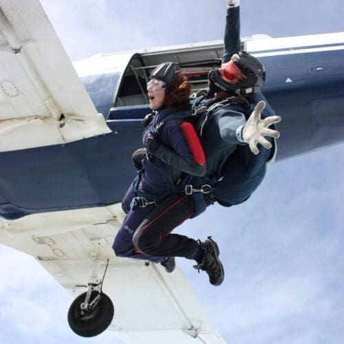 Two people strapped together for a tandem skydive have just jumped out of a small plane. The person with the parachute has their arms outstretched.