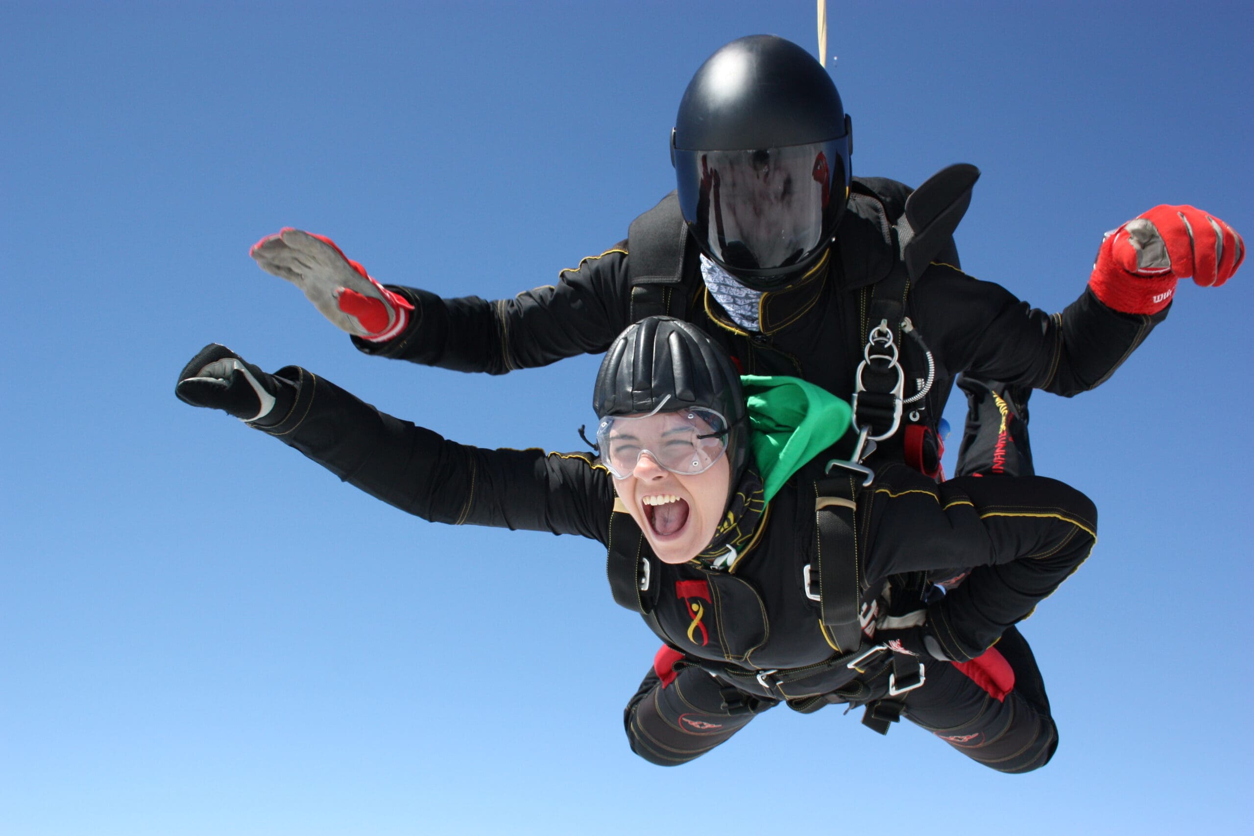 Close-up of tandem skydivers in freefall. The instructor at the back has a dark visor on their helmet. The person strapped to their front is wearing goggles and has their mouth open in an exhilarated yell.