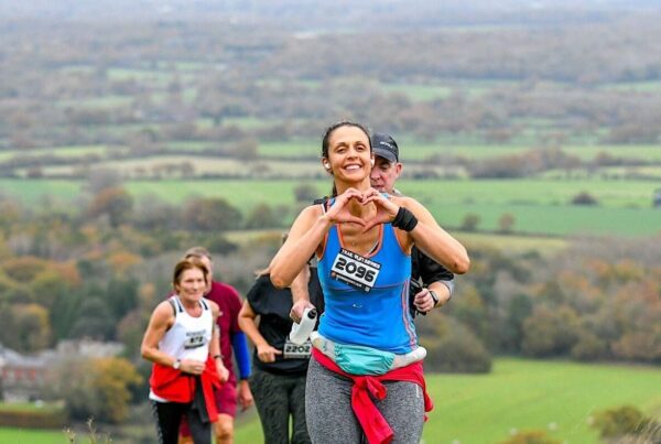 A group of five runners reach the summit of a hill in single file. They look tired but happy and determined. The lead runner is a woman who is smiling broadly and has her hands up in a heart shape gesture. Behind them is rolling green countryside.