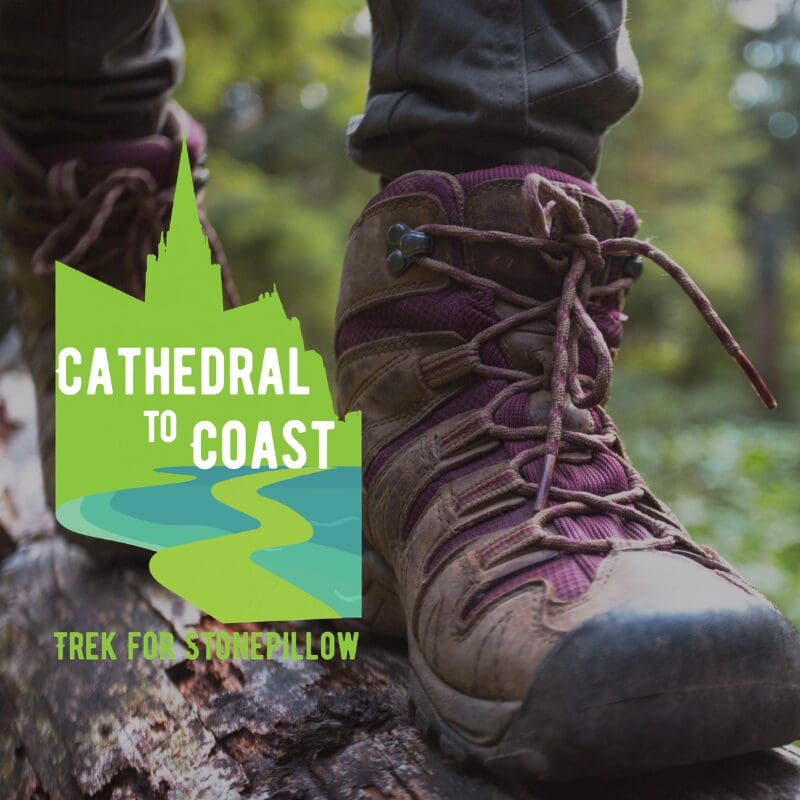 Close-up of someone's walking boots as they walk in the countryside. Logo for "Cathedral to Coast: Trek for Stonepillow" overlays the image