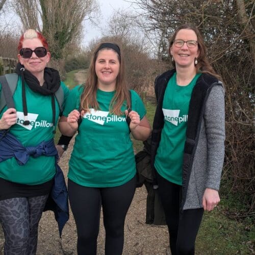 Three women dressed in walking gear and wearing Stonepillow branded green t-shirts are standing on a path in the countryside and smiling at the camera.