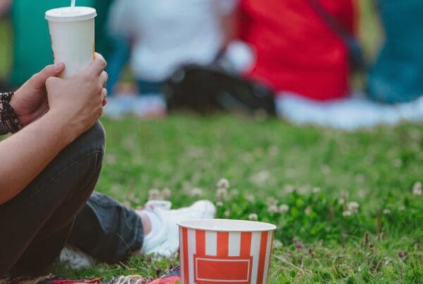 A person in jeans and white trainers is sitting on a picnic blanket on the grass holding a drink in a takeaway cup, with a popcorn bucket on the ground next to them. In the background an out-of-focus group of four people in bright colours are sitting in a row, as though watching a cinema screen