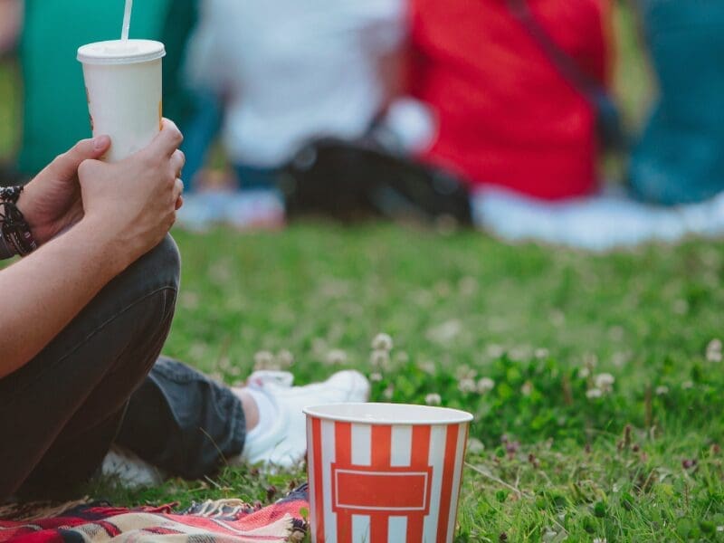 A person in jeans and white trainers is sitting on a picnic blanket on the grass holding a drink in a takeaway cup, with a popcorn bucket on the ground next to them. In the background an out-of-focus group of four people in bright colours are sitting in a row, as though watching a cinema screen