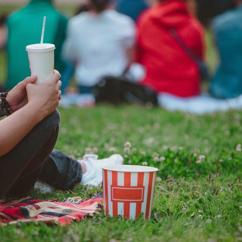 A person in jeans and white trainers is sitting on a picnic blanket on the grass holding a drink in a takeaway cup, with a popcorn bucket on the ground next to them. In the background an out-of-focus group of four people in bright colours are sitting in a row, as though watching a cinema screen
